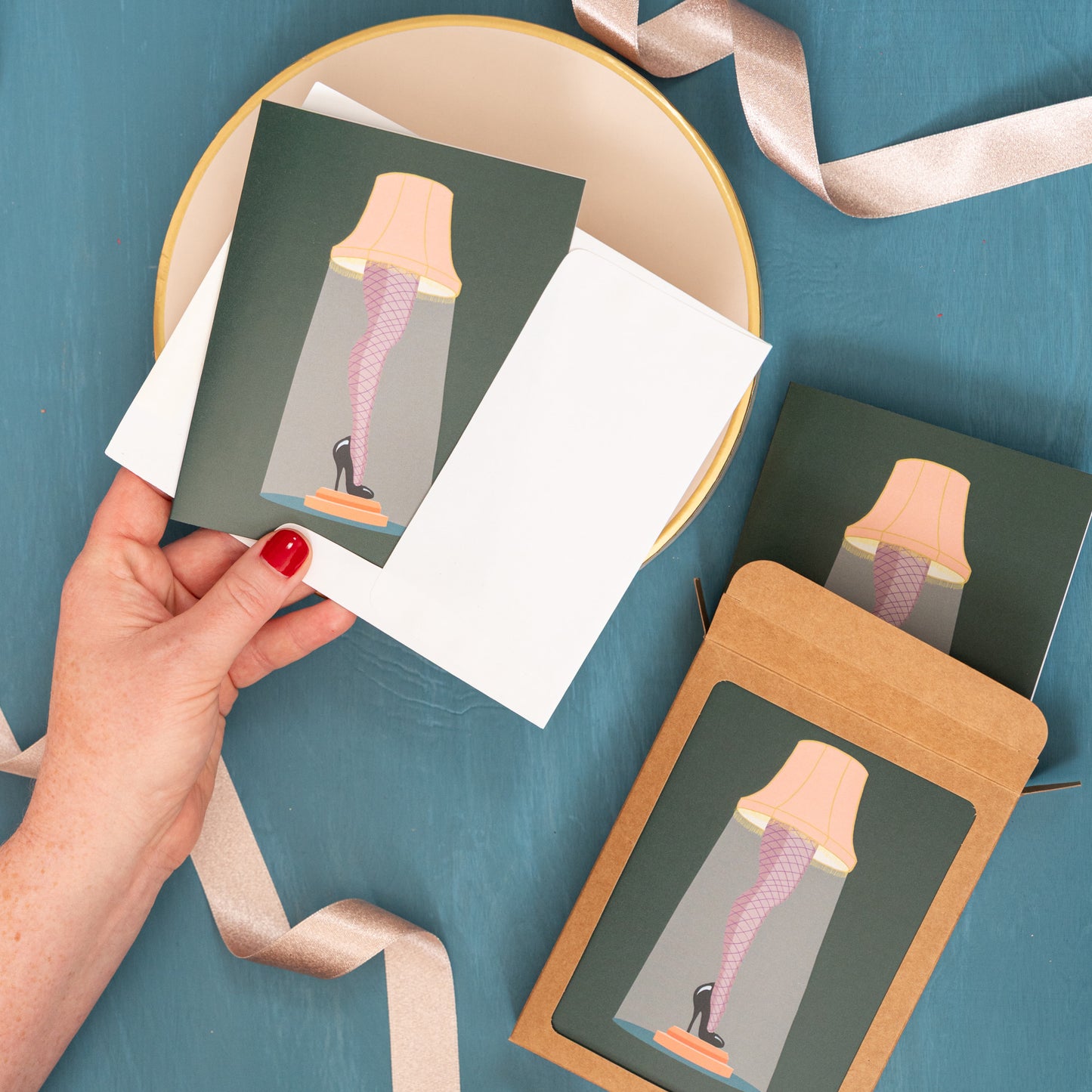 A hand holds a Christmas card with a leg lamp against a festive blue background with pink ribbons and a box of the same holiday card.