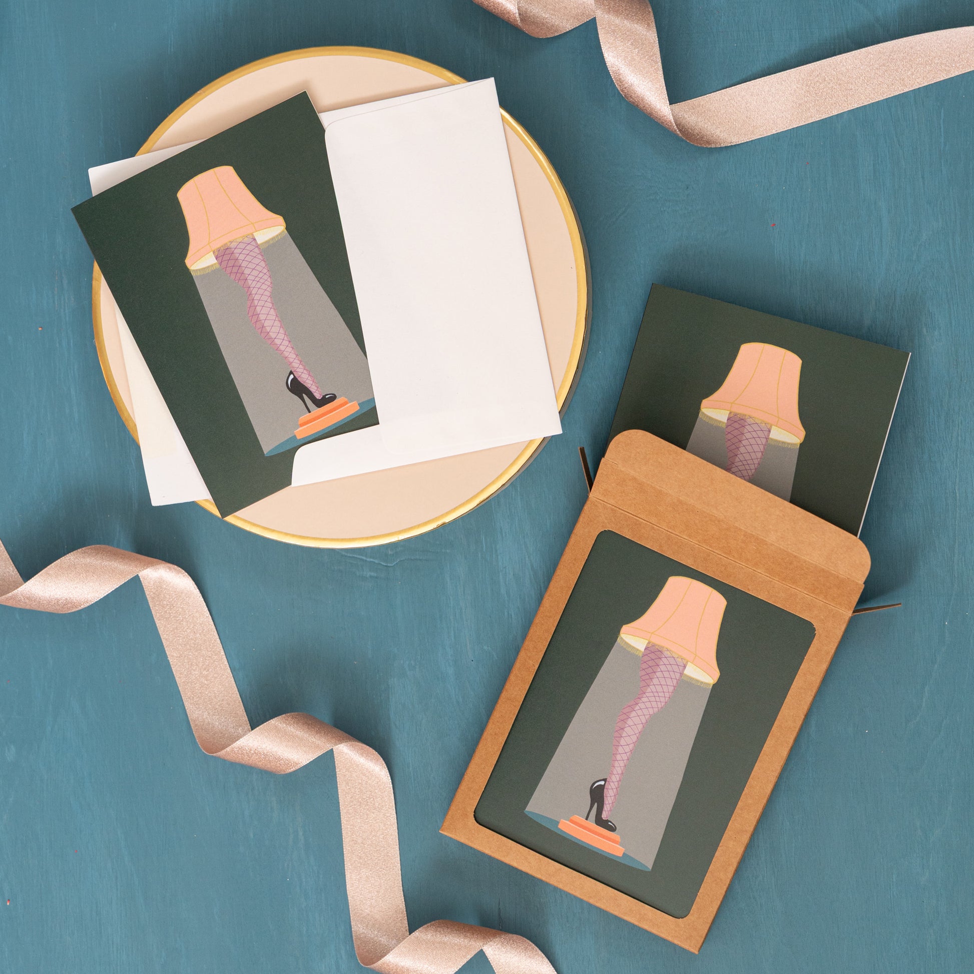 A Christmas card with a leg lamp appears against a festive blue background with pink ribbons and a box of the same holiday card.