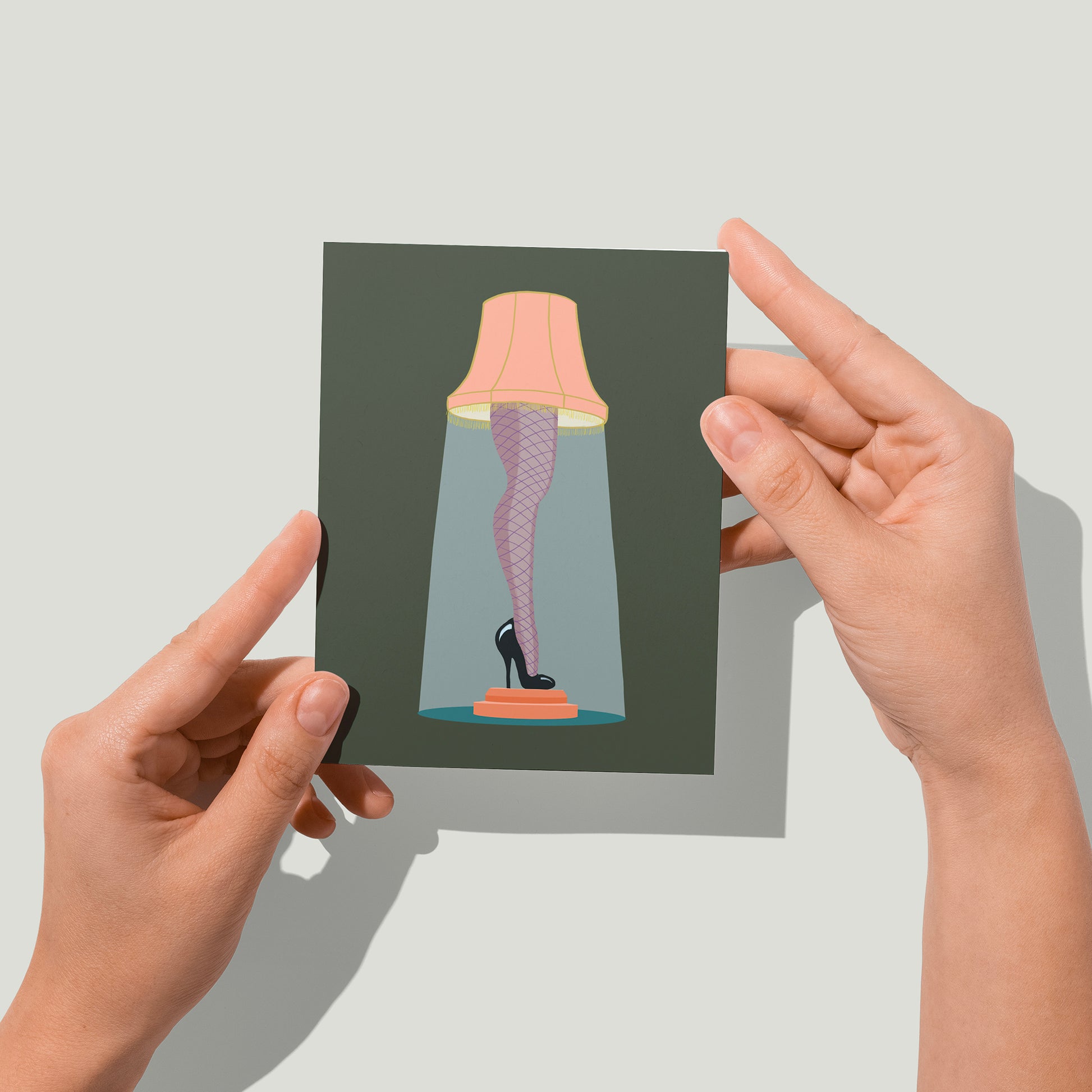 A dark green greeting card features an illustration of a lampshade atop a leg clad in fishnet stockings with a black high heel. A pair of hands holds the card up in front of the camera.