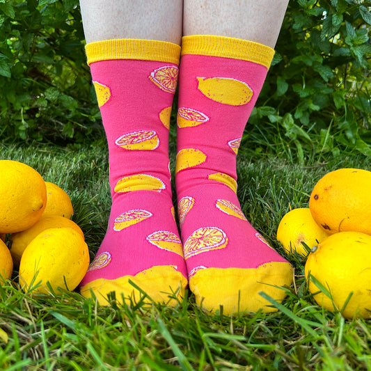 A person wears pink and yellow socks patterned with lemons. They are standing in the grass and lemons surround their feet.