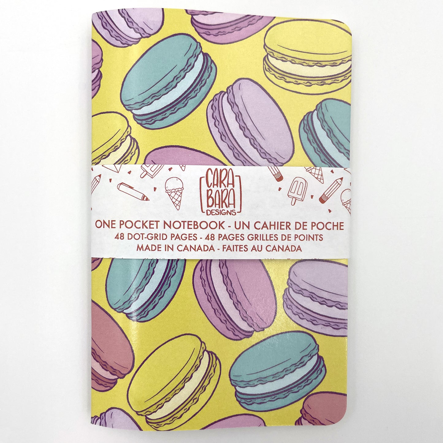 A small closed notebook has a cover that shows a rainbow of macarons against a yellow background. The notebook is bound with a Carabara Designs belly band indicating in both French and English that it is a pocket notebook containing 48 dot grid pages and is made in Canada.