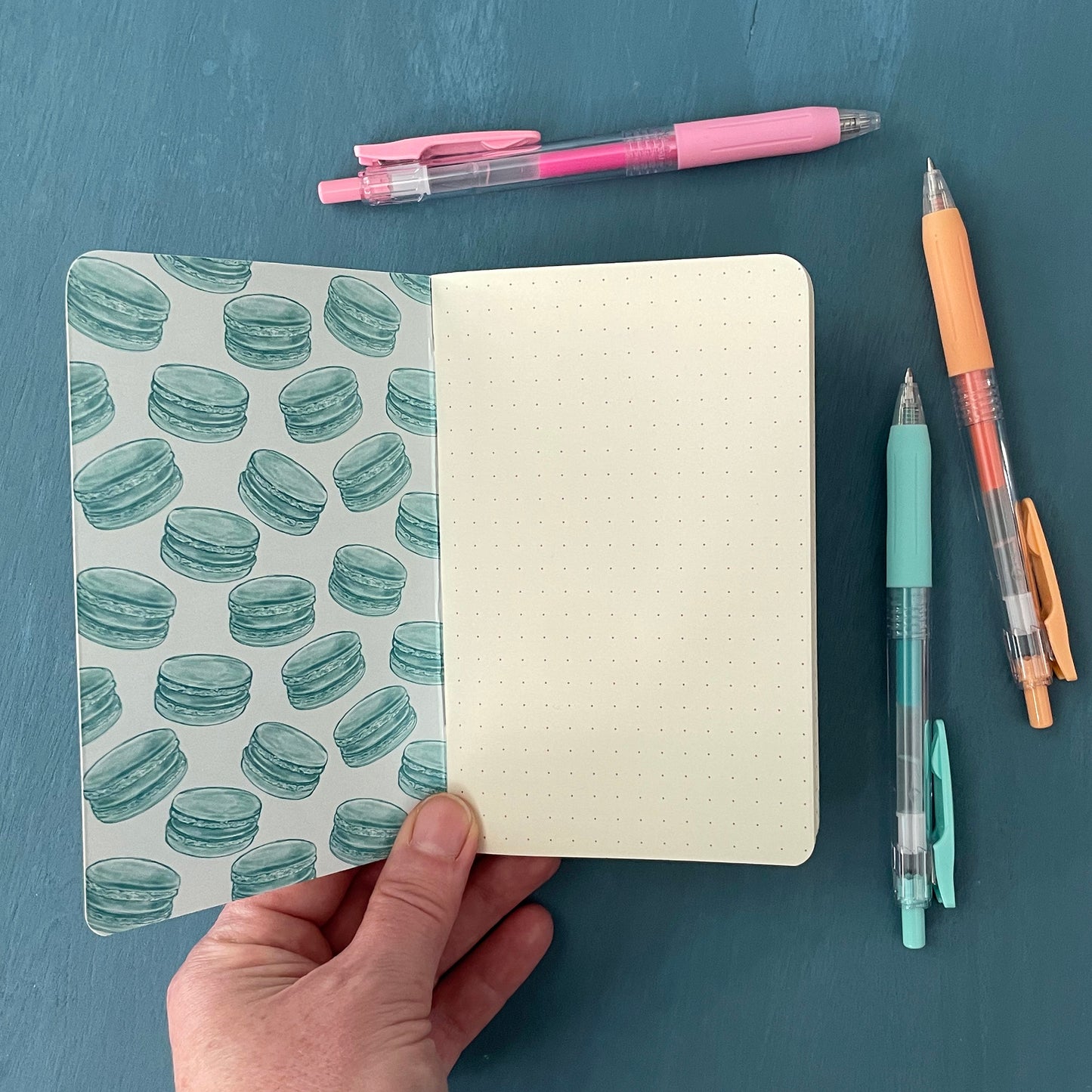 A pocket-sized notebook is open to its first page. The inner cover is patterned with pale blue macarons and the first page is covered in a deep grey-blue dot grid. The notebook sits on a blue background surrounded by pens.