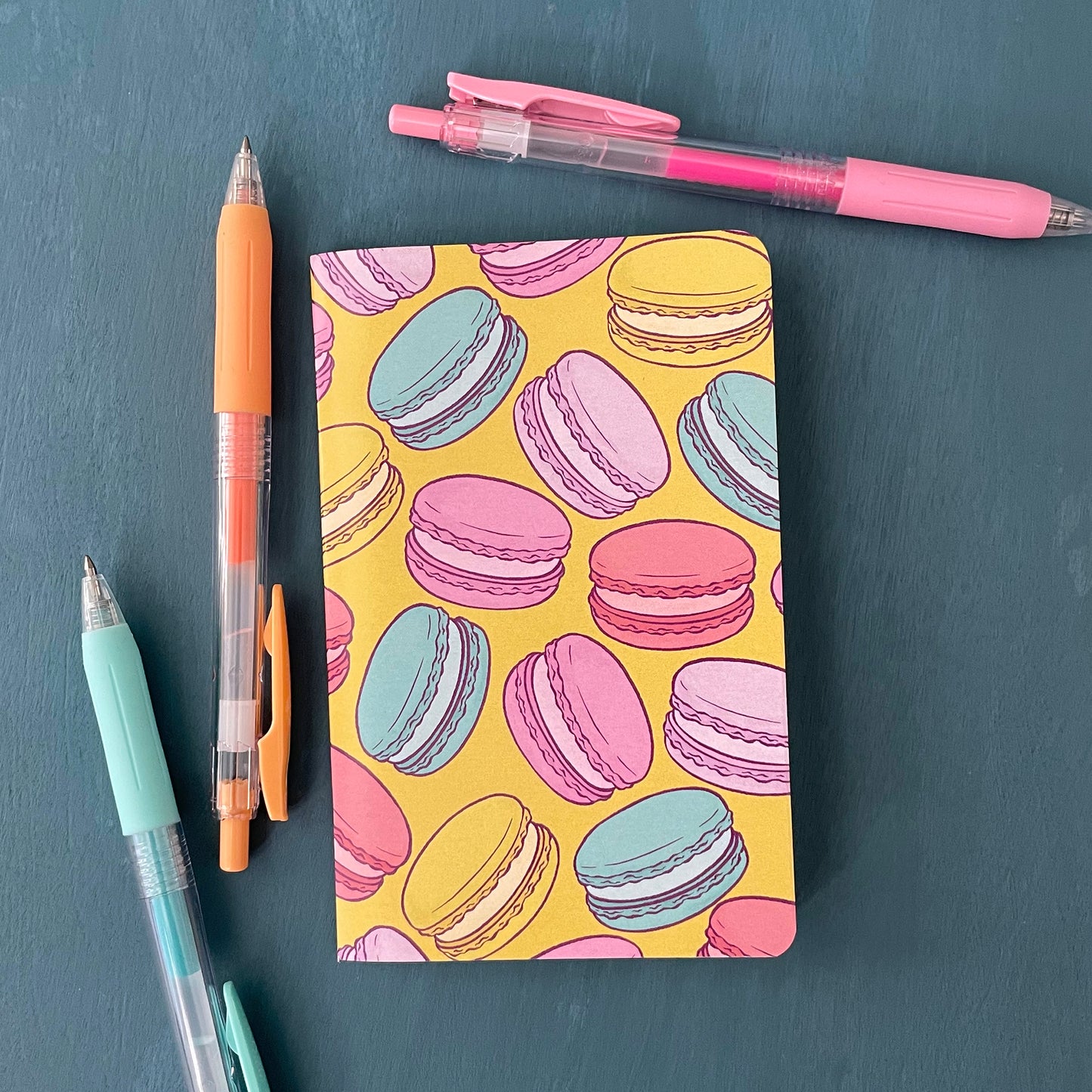 A pocket-sized notebook has a yellow cover patterned with French macarons. It sits on a blue background surrounded by pens.