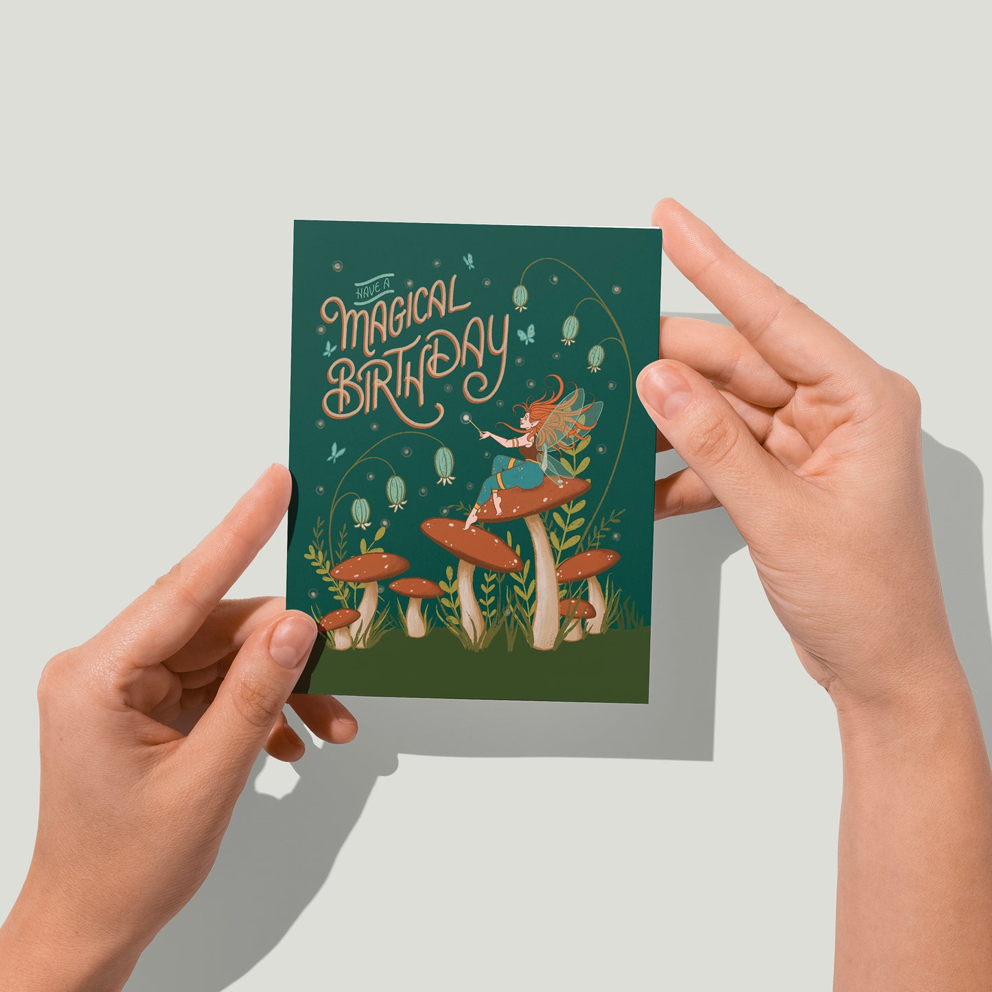 A dark green card says Have a Magical Birthday in blue letters. Below it on some grass are mushrooms, and a winged, red-haired faerie sits on one and looks up at the letters. Wildflowers appear behind the mushrooms. A pair of hands holds the card up in front of the camera.