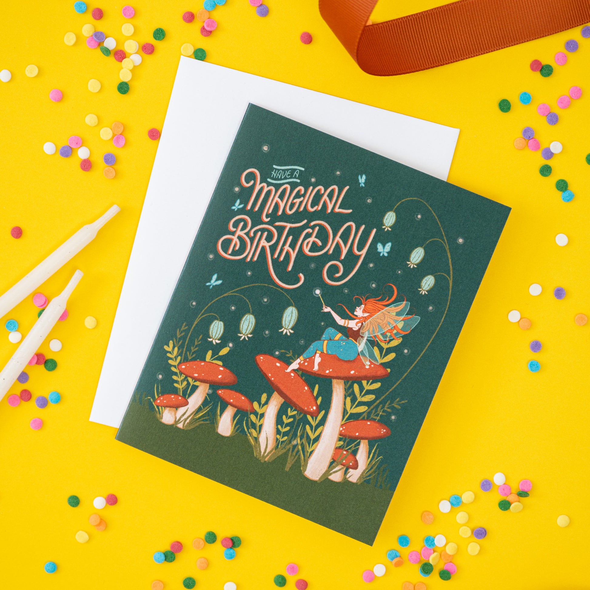 A dark green card shows a fairy, mushrooms, and the words 'have a magical birthday.' The card is on a yellow background with ribbon and confetti.