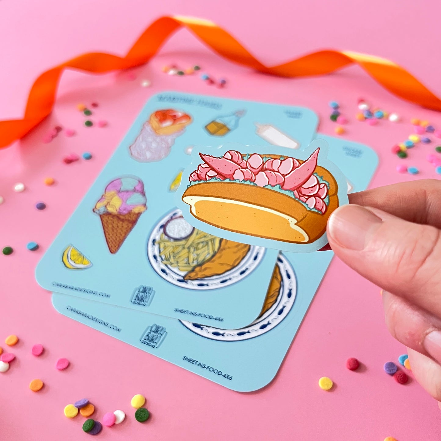 A lobster roll sticker is peeled from a sticker sheet full of Nova Scotia foods against a pink background.