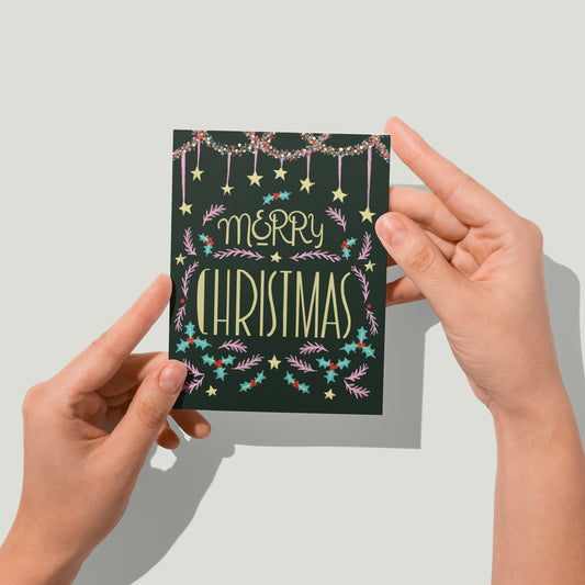Hands hold a dark Christmas card with hand lettering, starts and flourishes up to the camera.
