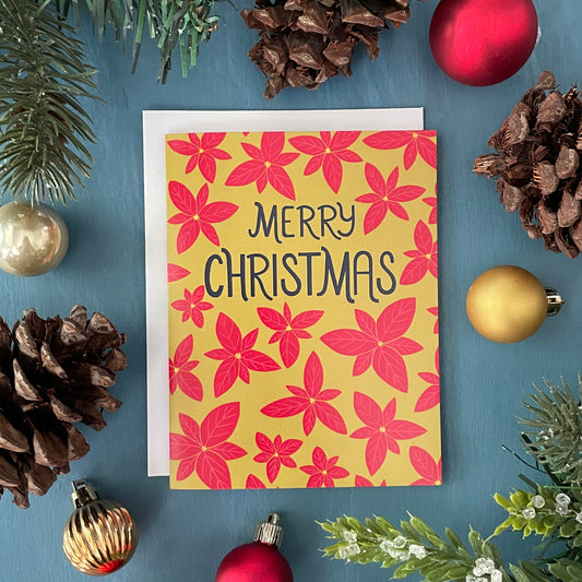 A bright green card features a pattern of bright red poinsettias and the words Merry Christmas in navy in the upper third. The card is surrounded by faux greenery, pinecones, and Christmas ornaments.