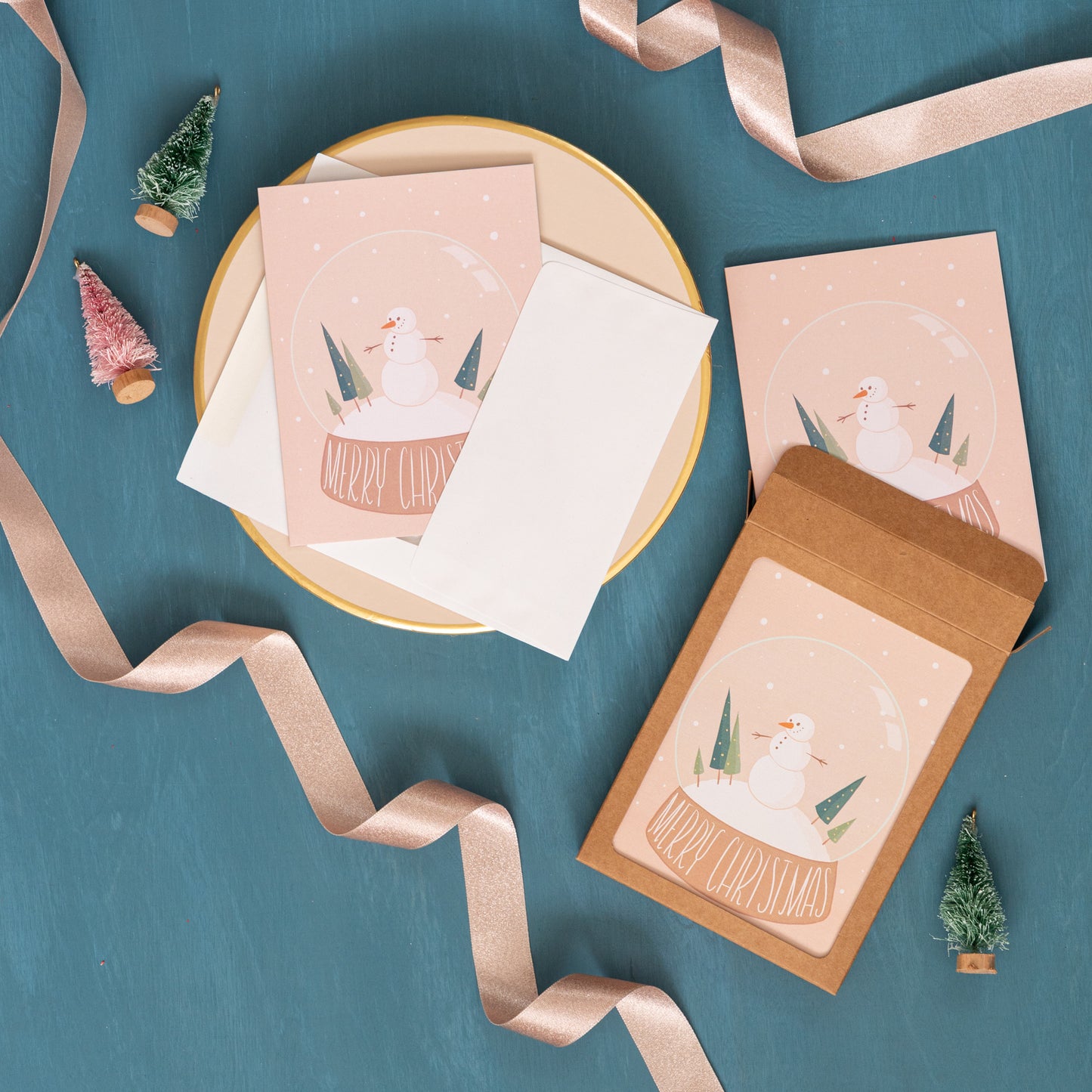 A snow globe Christmas card appears against a festive background of pink ribbon and bottle brush trees next to a box set of snowman holiday cards.