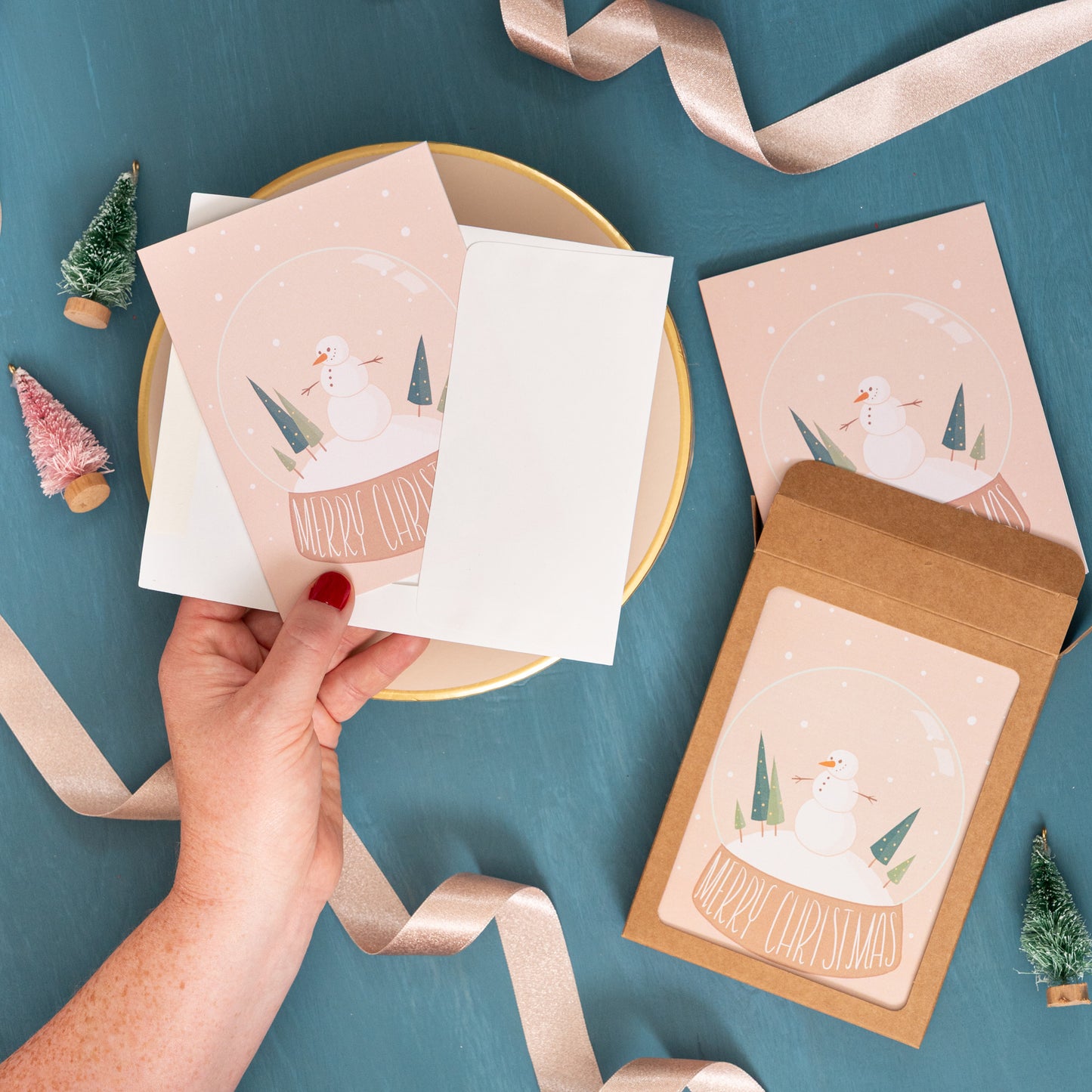 A hand holds a snow globe Christmas card against a festive background of pink ribbon and bottle brush trees.