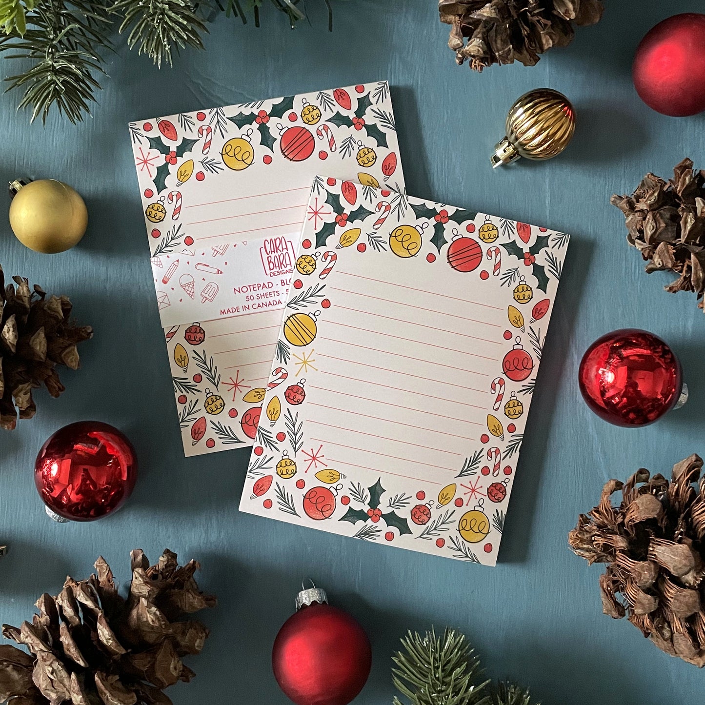 Two pale peach lined notepads featuring illustrations of holly, ornaments, Christmas lights, candy canes, and sprigs of greenery are sitting among holiday ornaments and greenery. One notepad is packaged with a belly band saying that it is 50 sheets and made in Canada.