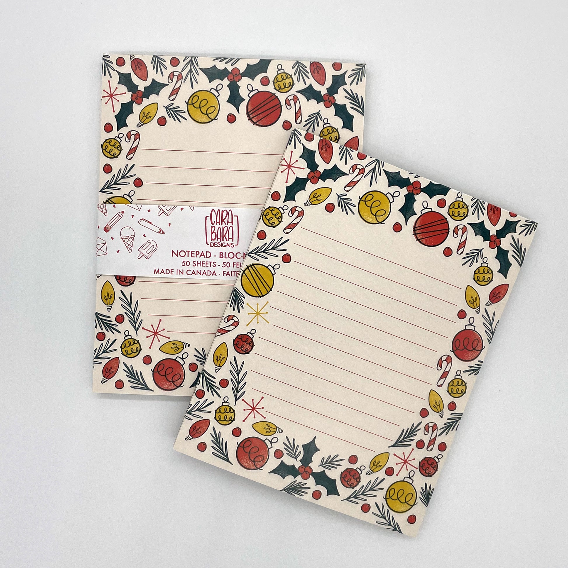 Two pale peach lined notepads featuring illustrations of holly, ornaments, Christmas lights, candy canes, and sprigs of greenery are sitting on a white background. One notepad is packaged with a belly band saying that it is 50 sheets and made in Canada.