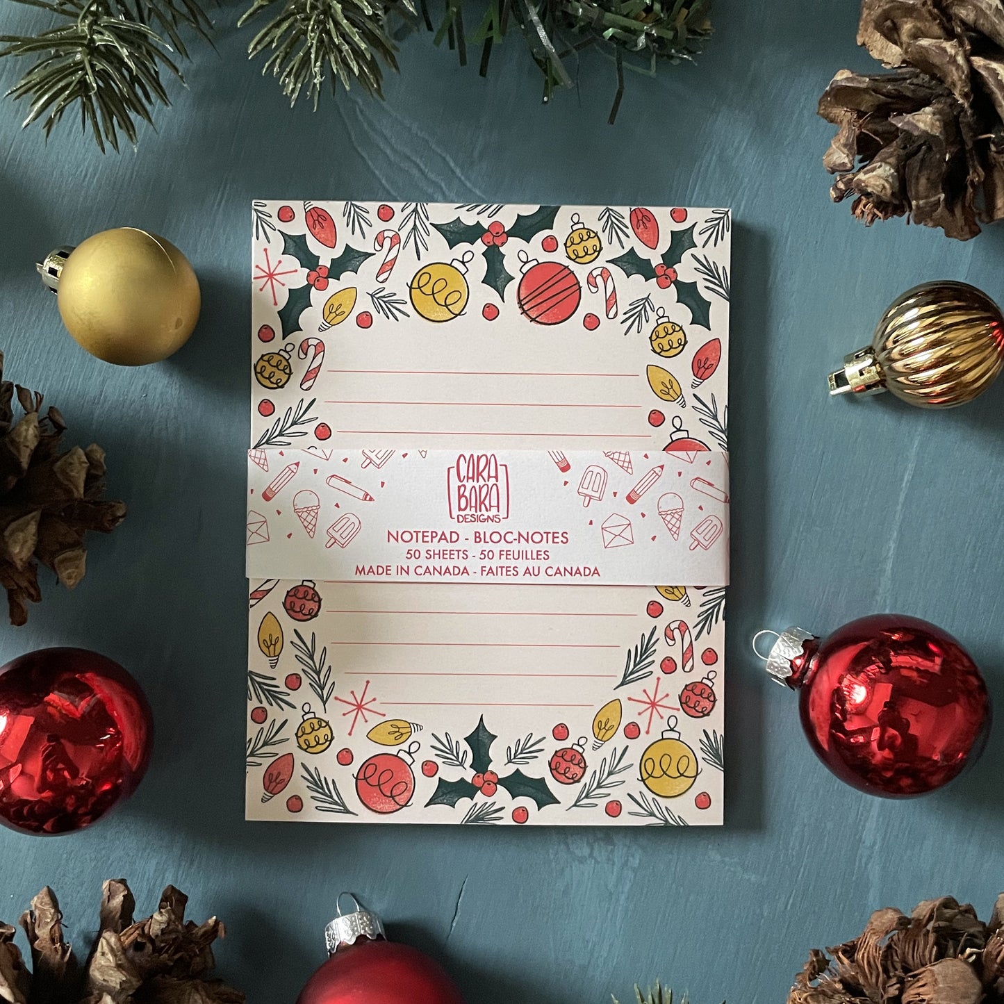 A pale peach lined notepad featuring illustrations of holly, ornaments, Christmas lights, candy canes, and sprigs of greenery is sitting among holiday ornaments and greenery. The notebook is packaged with a band saying that it is 50 pages and made in Canada.