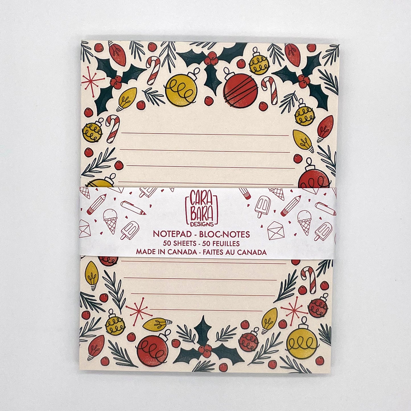 A pale peach lined notepad features illustrations of holly, ornaments, christmas lights, candy canes, and pine sprigs. The notebook is packaged with a band saying that it is 50 pages and made in Canada.