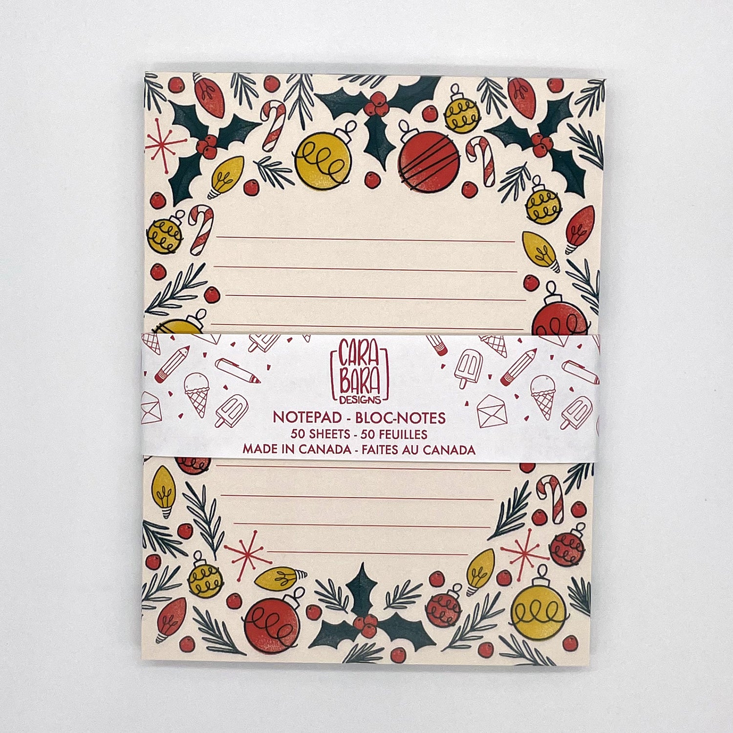 A pale peach lined notepad features illustrations of holly, ornaments, christmas lights, candy canes, and pine sprigs. The notebook is packaged with a band saying that it is 50 pages and made in Canada.