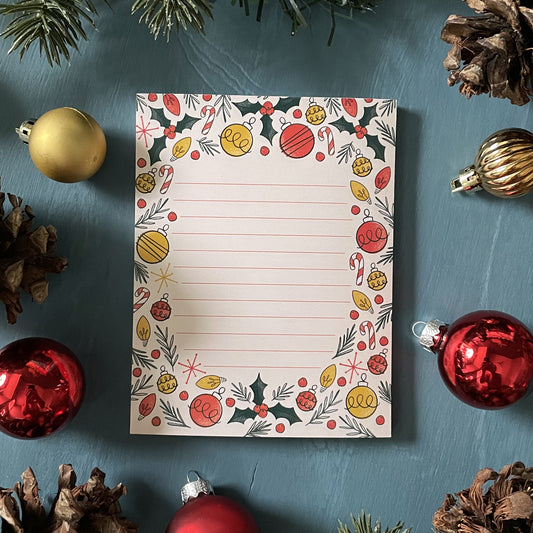 A pale peach lined notepad featuring illustrations of holly, ornaments, Christmas lights, candy canes, and sprigs of greenery is sitting among holiday ornaments and greenery.