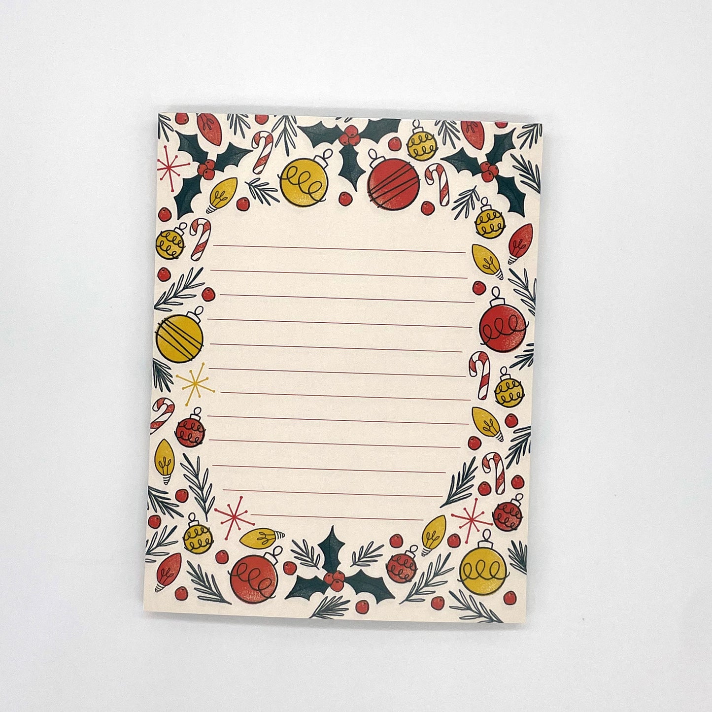 A pale peach lined notepad features illustrations of holly, ornaments, christmas lights, candy canes, and pine sprigs.