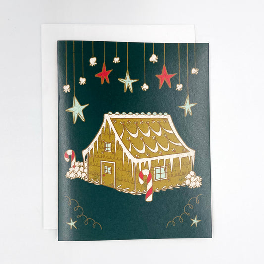 A dark green greeting card features hanging stars and a frosted gingerbread house among popcorn snowbanks. The card is on a white envelope on a white background.