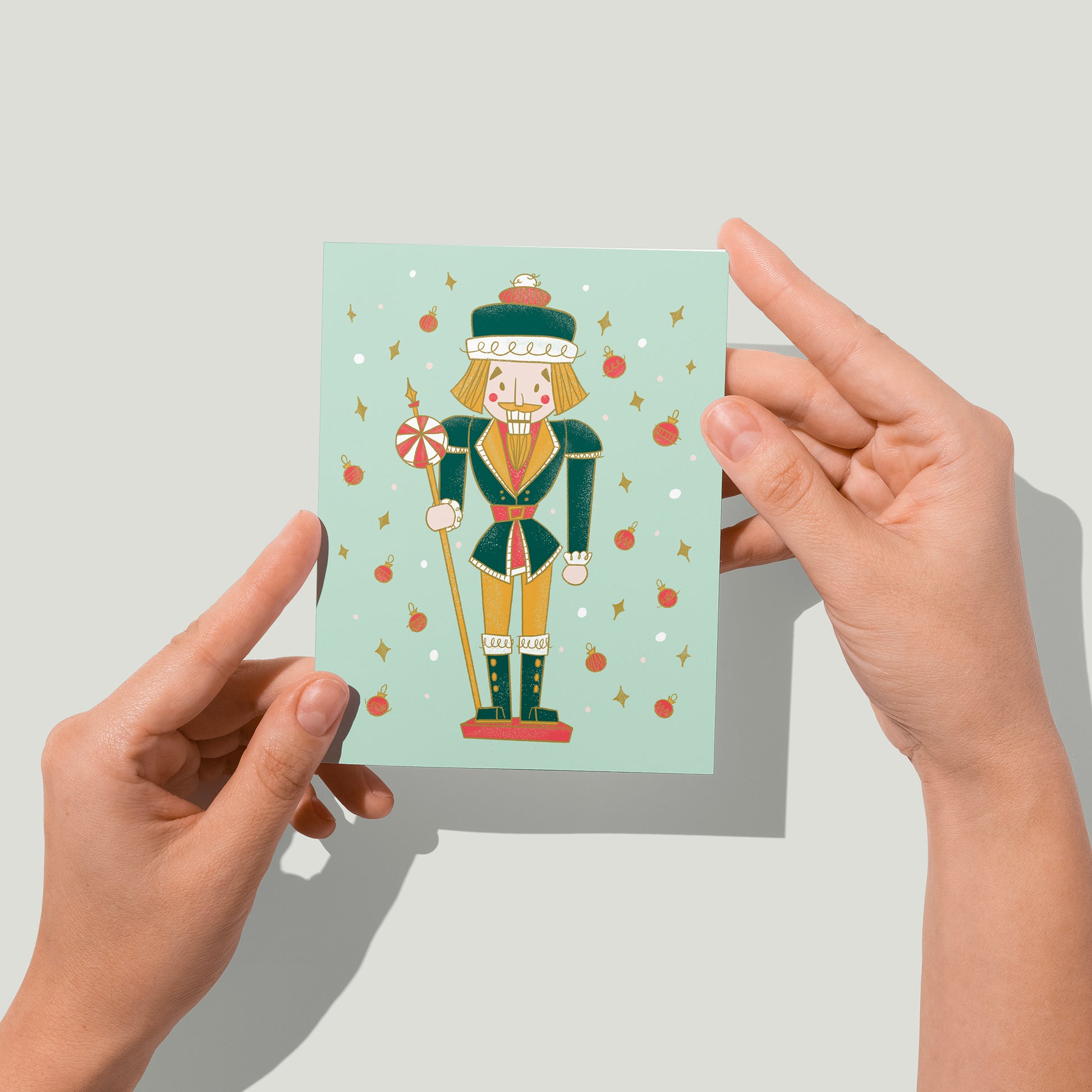 A mint card features a midcentury-style nutcracker in green and yellow, flanked by dots, yellow diamonds and red baubles. A pair of hands holds the card up in front of the camera.