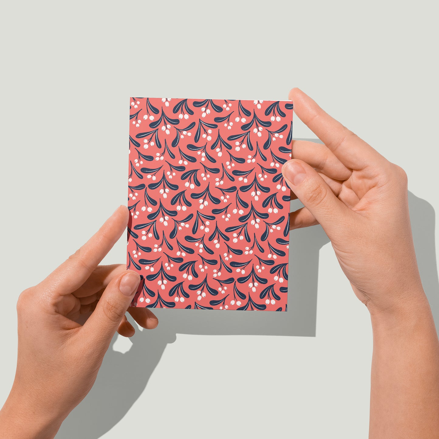 A red greeting card shows a blue and white botanical mistletoe pattern. A pair of hands holds the card up in front of the camera.