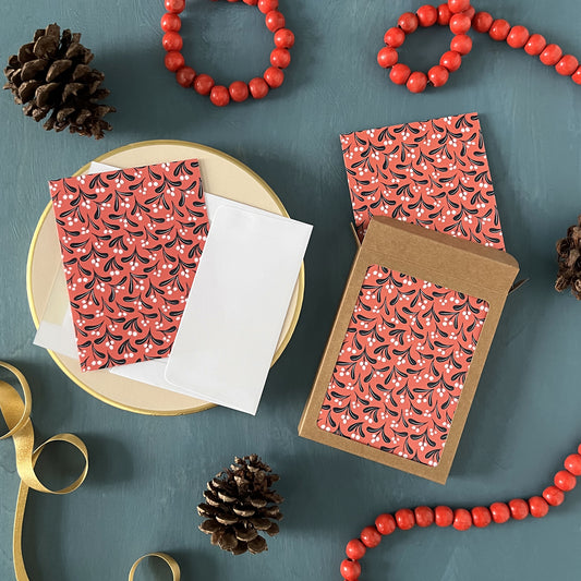 Brightly coloured mistletoe patterned Christmas greeting cards appear on a festive blue background with red beads, gold ribbon, and pinecones.