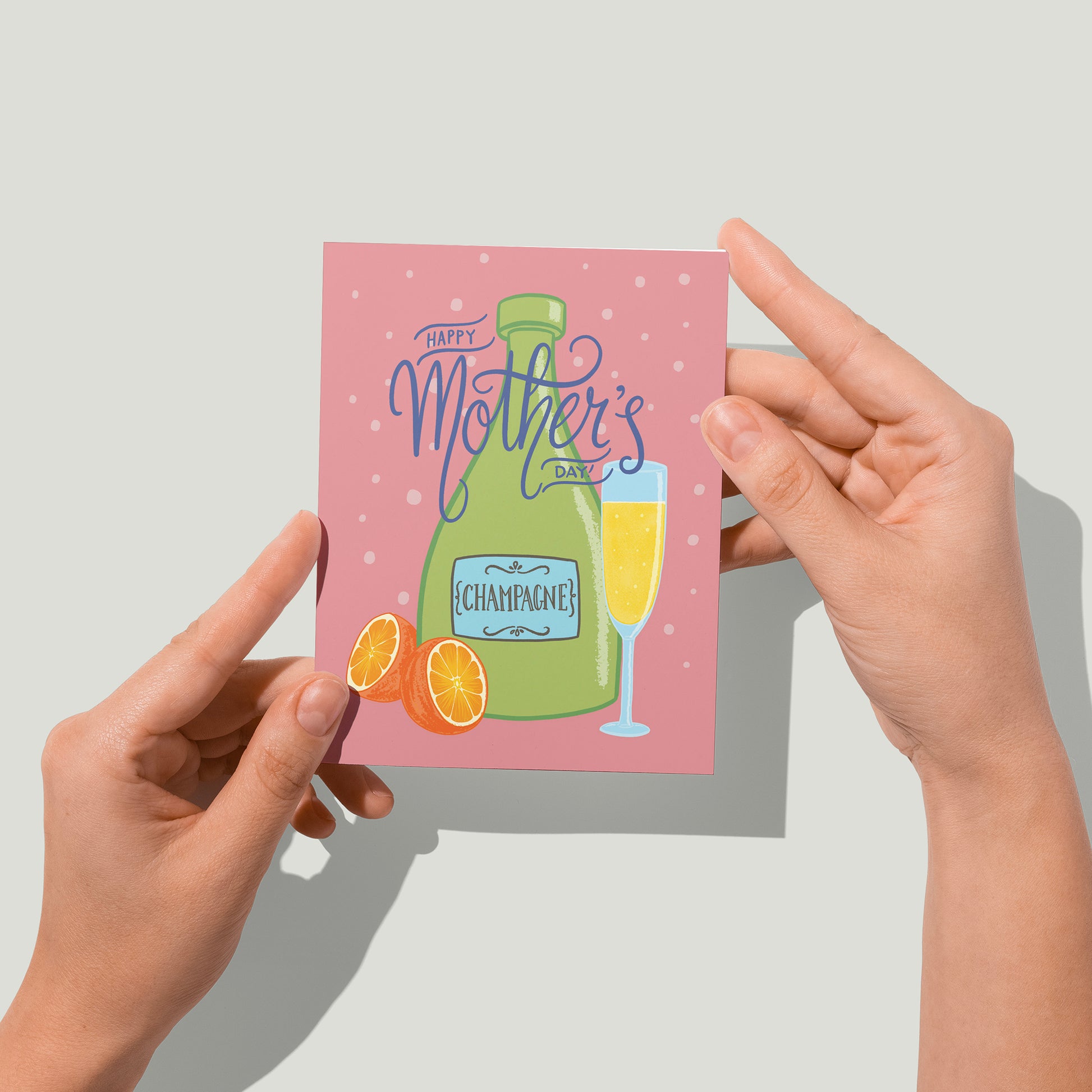 A pink greeting card reads Happy Mother's Day in blue hand lettering on top of an illustration of a green bottle of champagne, an orange cut in half, and a full champagne flute. A pair of hands holds the card up in front of the camera.