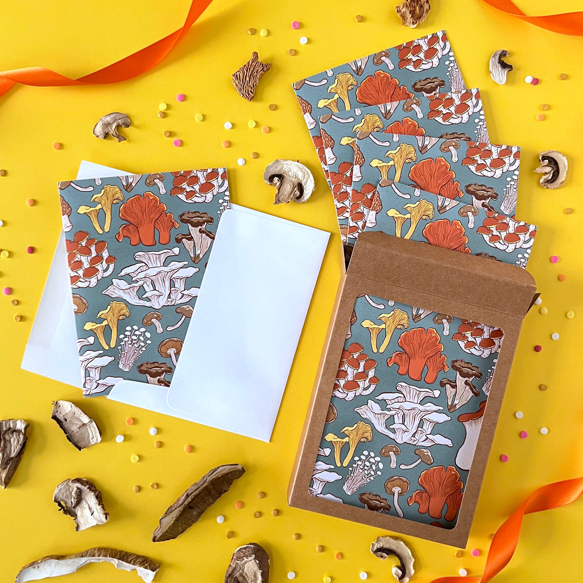 A box of greeting cards patterned with mushrooms spills onto yellow with confetti and dried mushrooms.