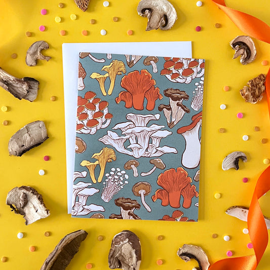 A greeting card patterned with a variety of mushrooms is on yellow with confetti, ribbon and dried mushrooms.