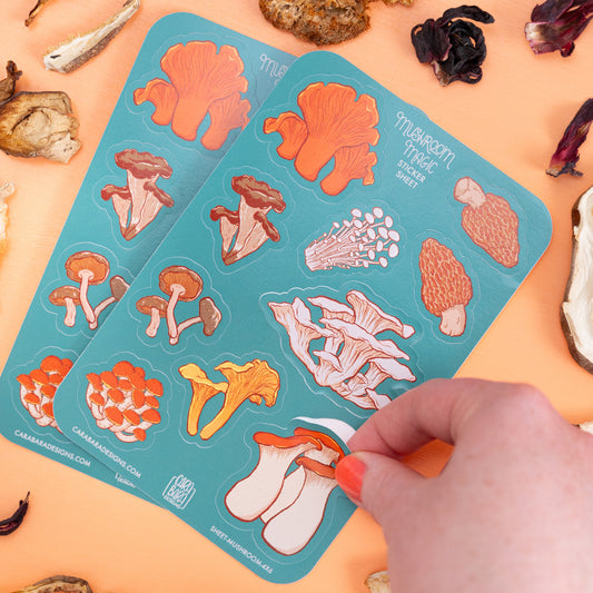 Two teal sticker sheets with a variety of mushroom stickers are on an orange background with mushrooms and ribbon. A hand peels off the king oyster mushroom sticker.
