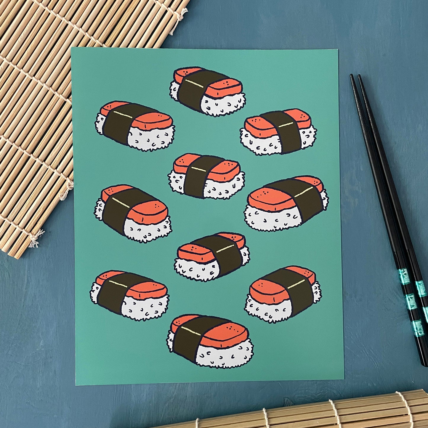 An art print shows a cluster of musubi-style sushi on a turquoise background. The art print sits on a sushi mat next to chopsticks.