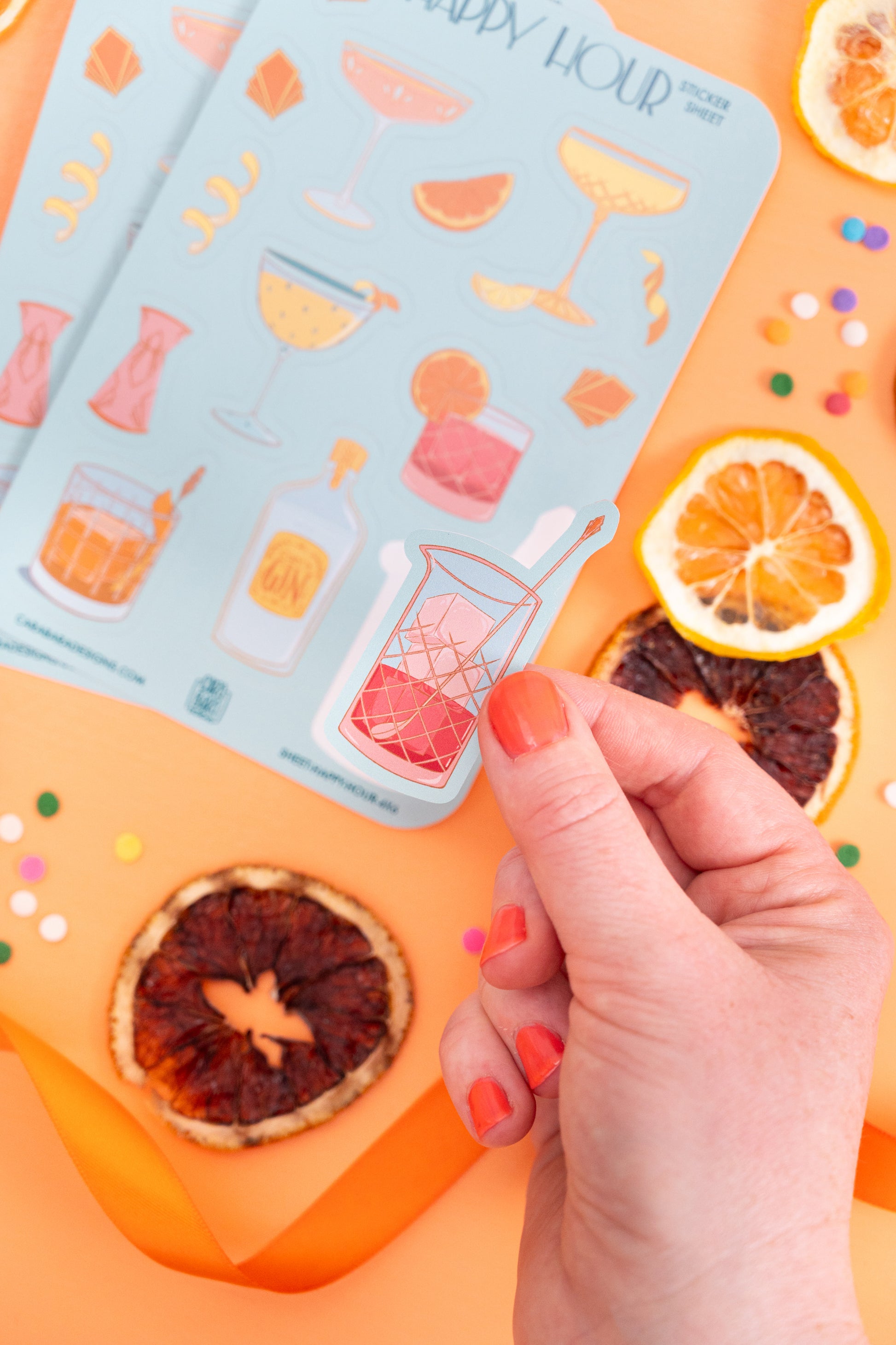 Two art deco cocktail-themed sticker sheets are on an orange background with citrus slices. A hand lifts the negroni mixing glass sticker off the front sheet.