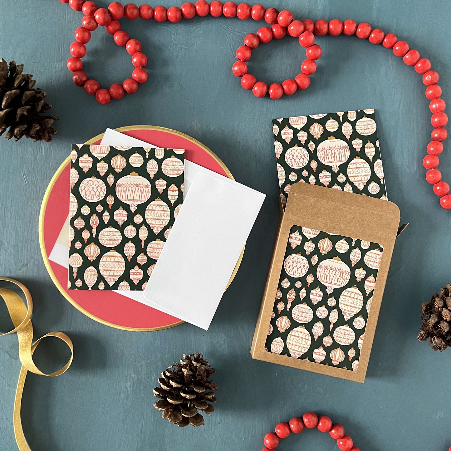 A dark green holiday greeting card patterned with elaborate Christmas ornamentsappears on a festive blue background with red beads and pinecones.