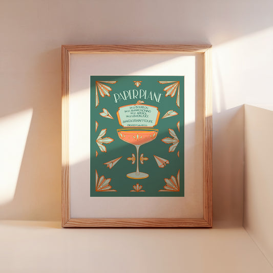 An art deco illustrated paper plane cocktail recipe leans framed against a wall.