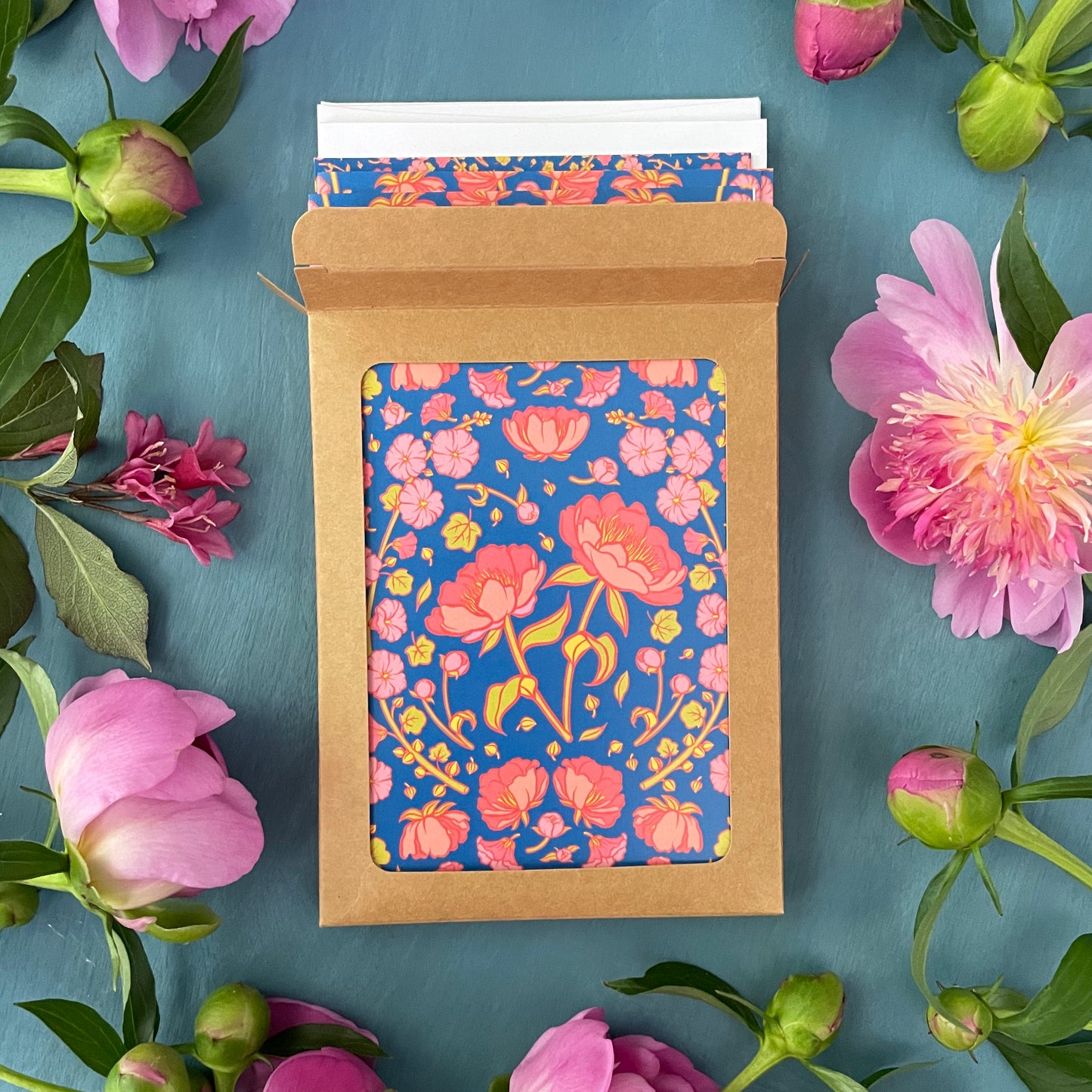 A blue greeting card features a symmetrical pattern of pink peony blossoms and pink hollyhock stalks. Peony cards spill out of an open kraft paper box surrounded by fresh pink peonies.