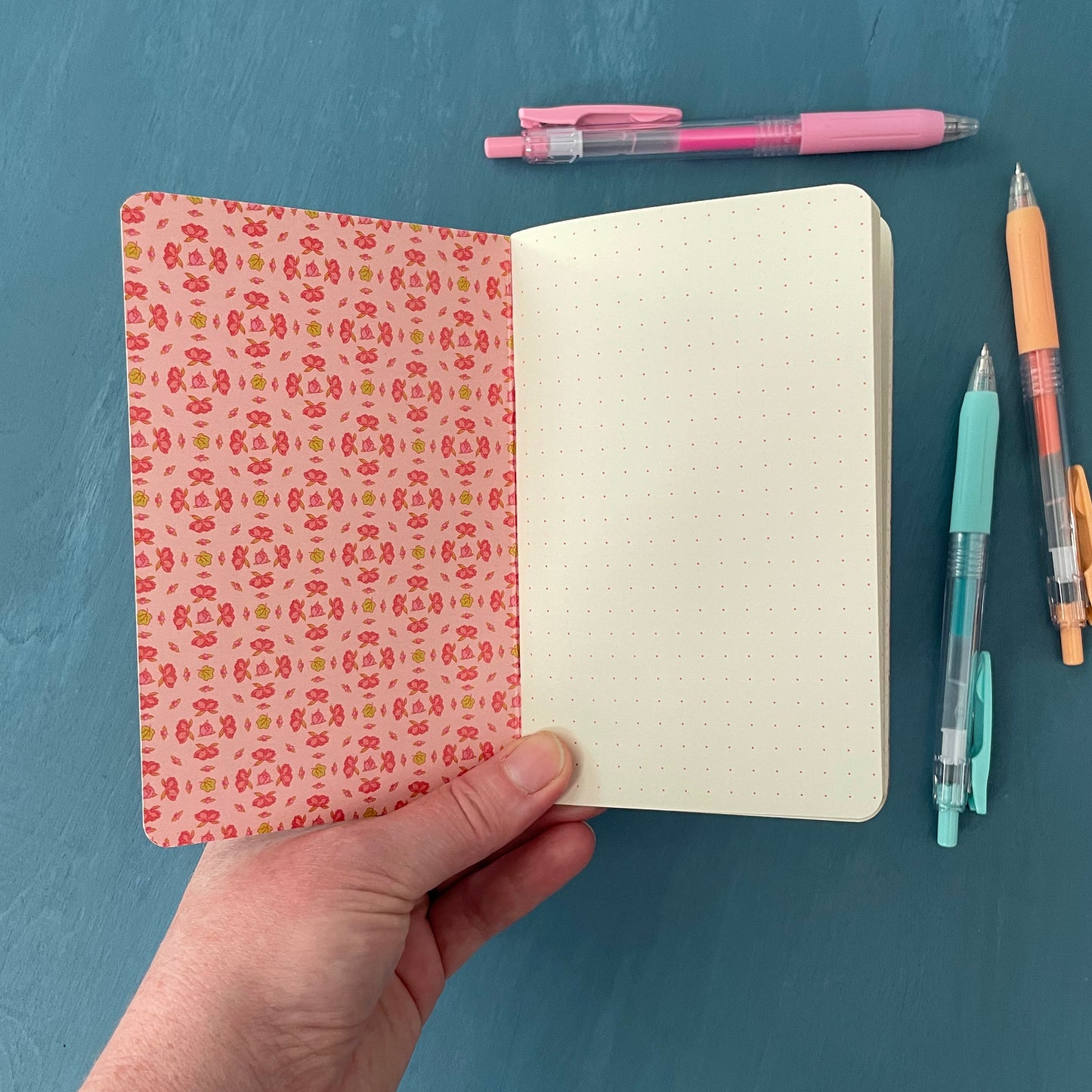 A pocket notebook is open to its first page. The inside cover is pink and patterned with a geometric arrangement of pink peony blossoms. The first page of the notebook is creamy paper stock covered with a pink dot grid. The notebook is on a blue background surrounded by pens.