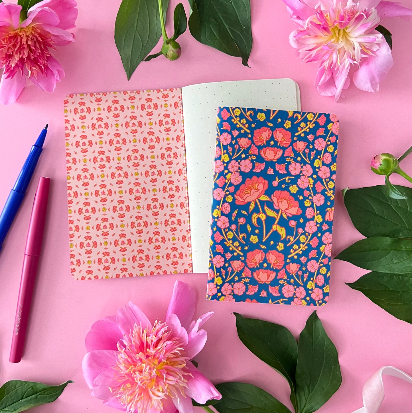A blue notebook with peonies on the cover is on top of an open dot grid notebook on pink with peonies around it.