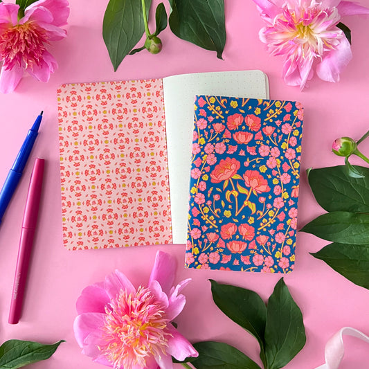 A blue notebook with peonies on the cover is on top of an open dot grid notebook on pink with peonies around it.
