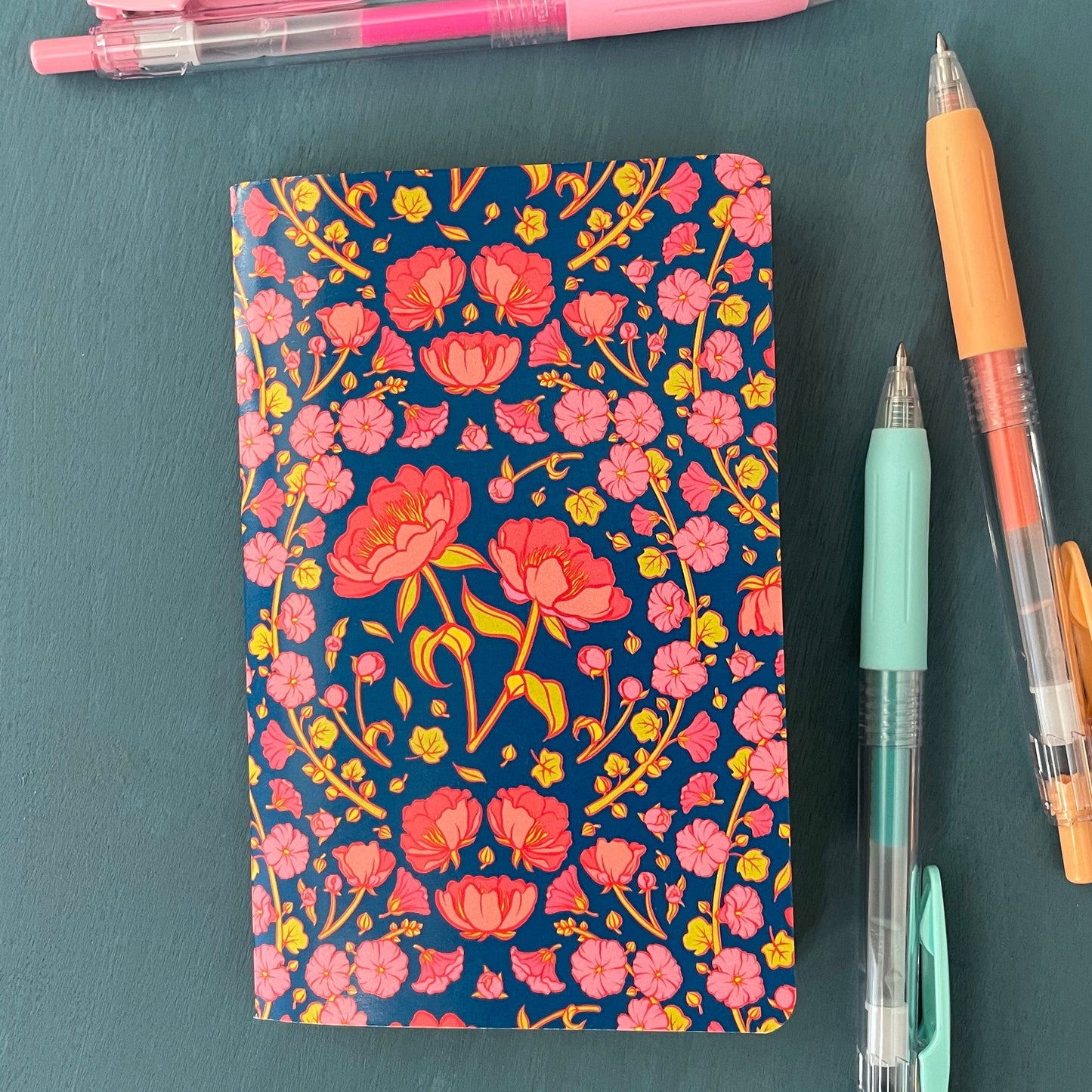 A blue floral notebook is covered in pink peonies and hollyhocks. It is surrounded by three pens.