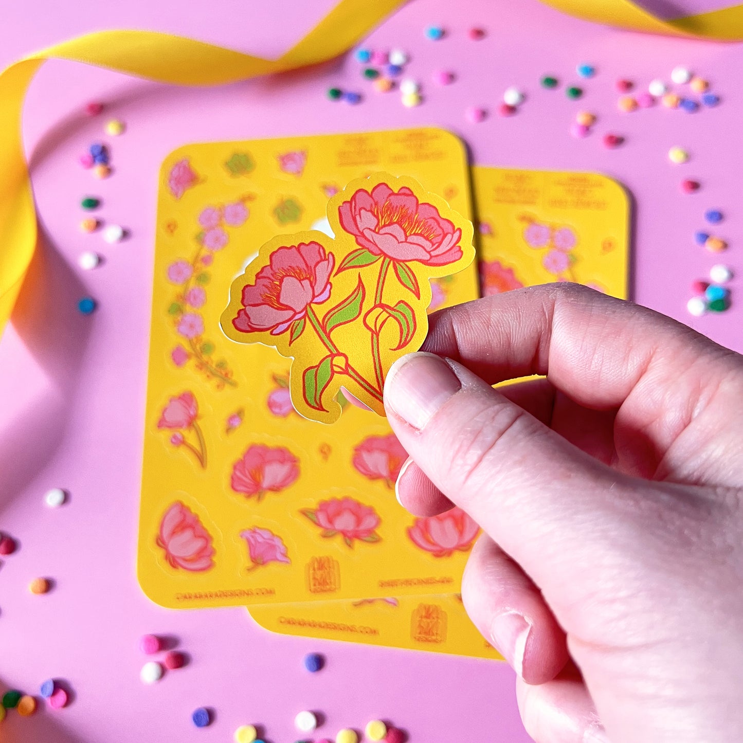 Two yellow sticker sheets featuring pink peony and pink hollyhock stickers are on a pink background with confetti and ribbon. A hand holds up a peony sticker to the camera.