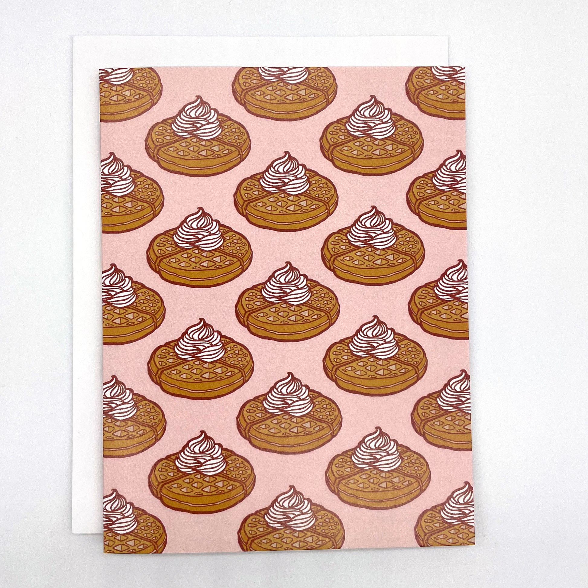 A pink greeting card patterned with Belgian waffles topped with whipped cream appears against a white envelope on a white background.