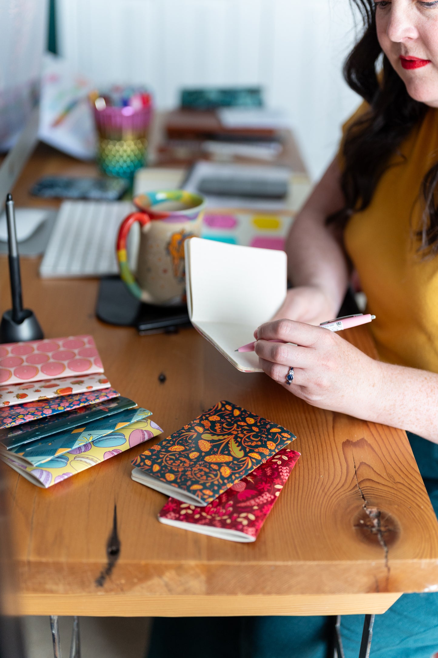 A woman writes in a small notebook at a desk. She is surrounded by colourful pocket notebooks.