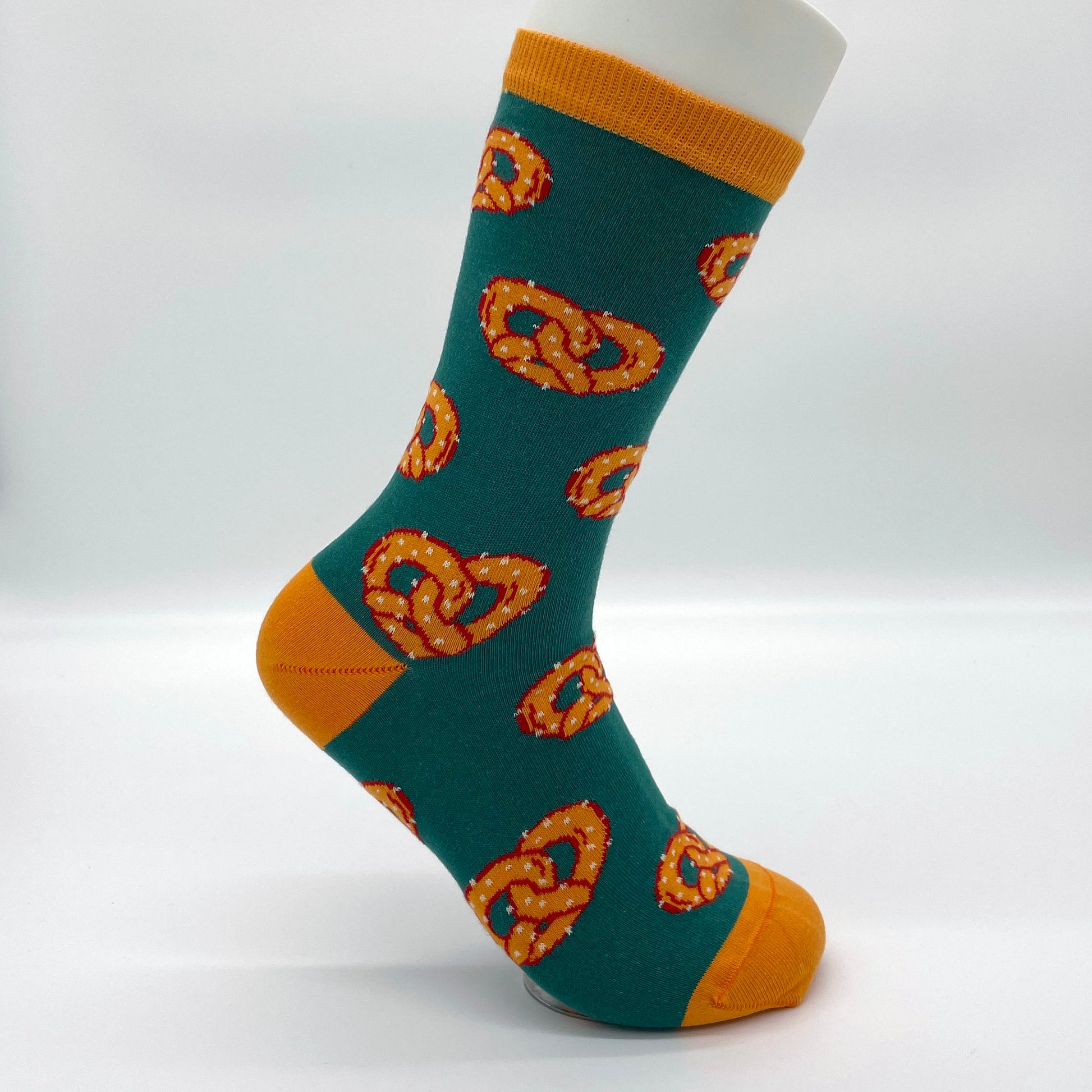 A white sock mannequin sports a green sock patterned with golden-orange salted soft pretzels. The welt, heel and toe of the sock are orange.