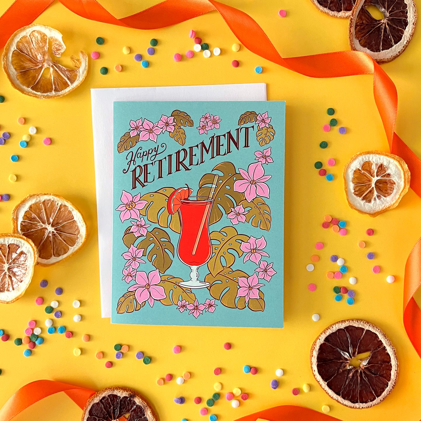 A blue retirement card with a hurricane cocktail, orchids and leaves is on yellow with dried citrus and confetti.