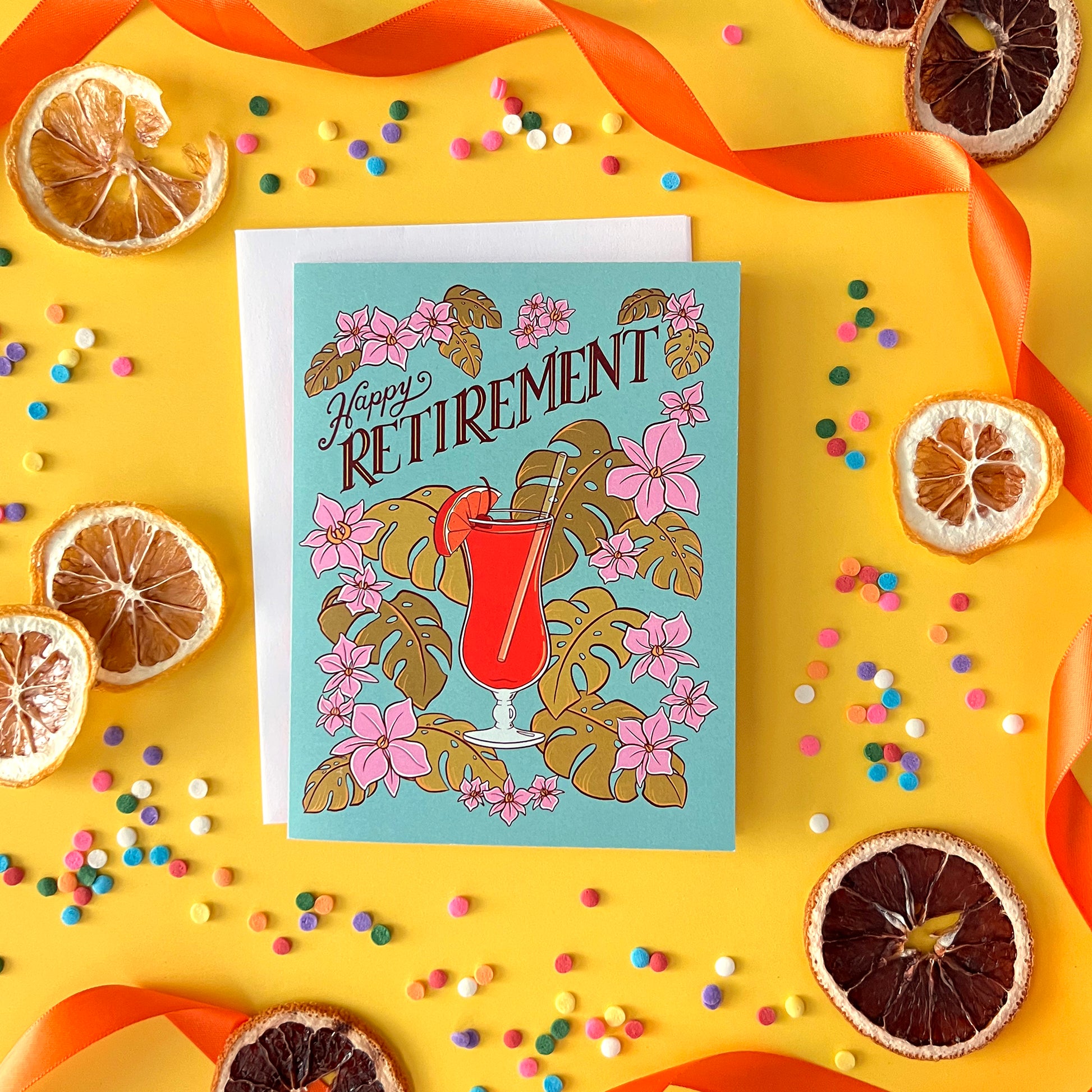 A blue retirement card with a hurricane cocktail, orchids and leaves is on yellow with dried citrus and confetti.