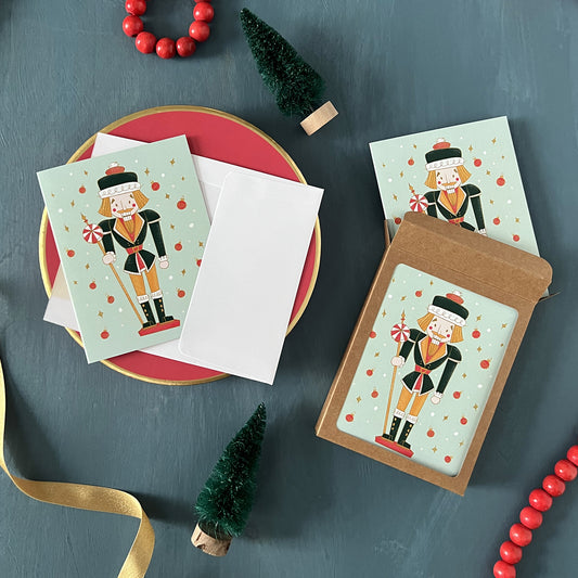 Mint green holiday greeting cards with retro nutcrackers appear on a festive blue background with red beads, green ribbon, and bottle brush trees.