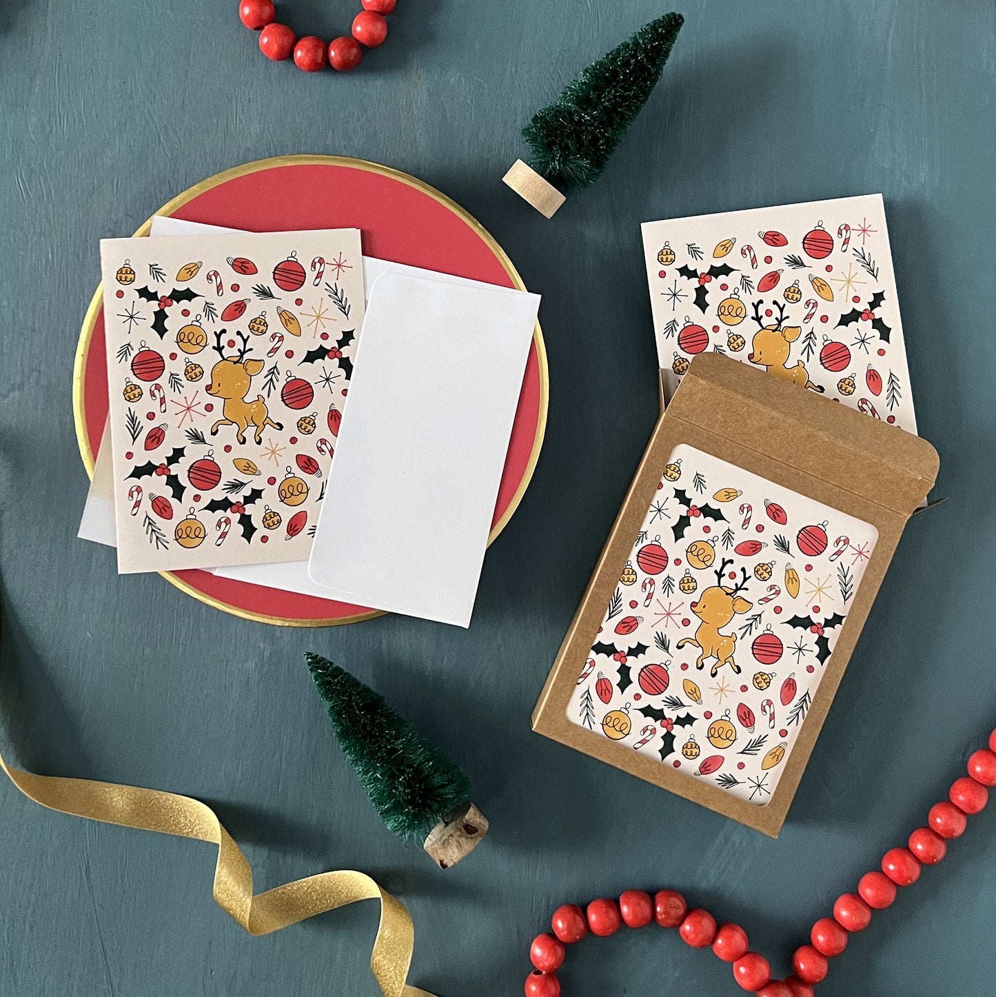 Holiday greeting cards with cute retro reindeer, ornaments, holly and Christmas lights appear on a festive blue background with red beads, green ribbon, and bottle brush trees.
