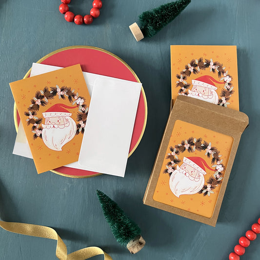 Yellow Holiday greeting cards with a cute retro Santa Claus head in a wreath appear on a festive blue background with red beads, green ribbon, and bottle brush trees.