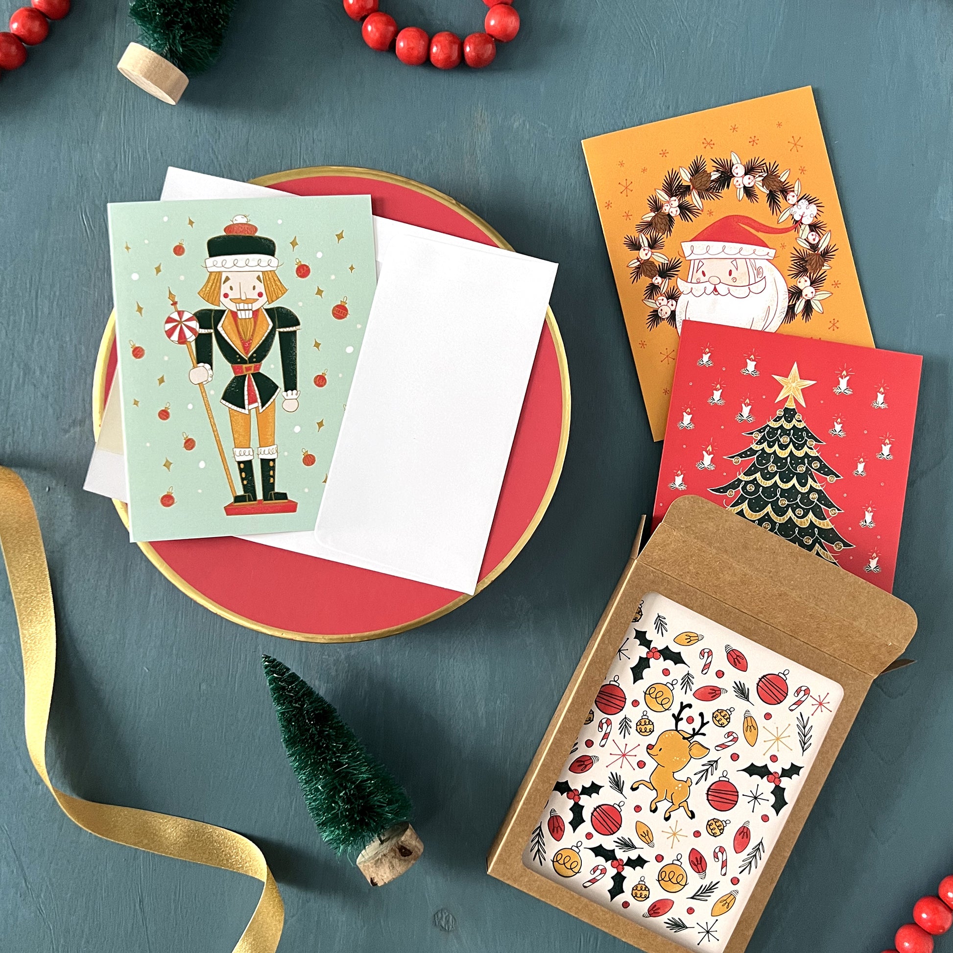 A boxed collection of 4 retro Christmas card designs appears against a festive blue background with bottle brush trees, red beads, and gold ribbon.