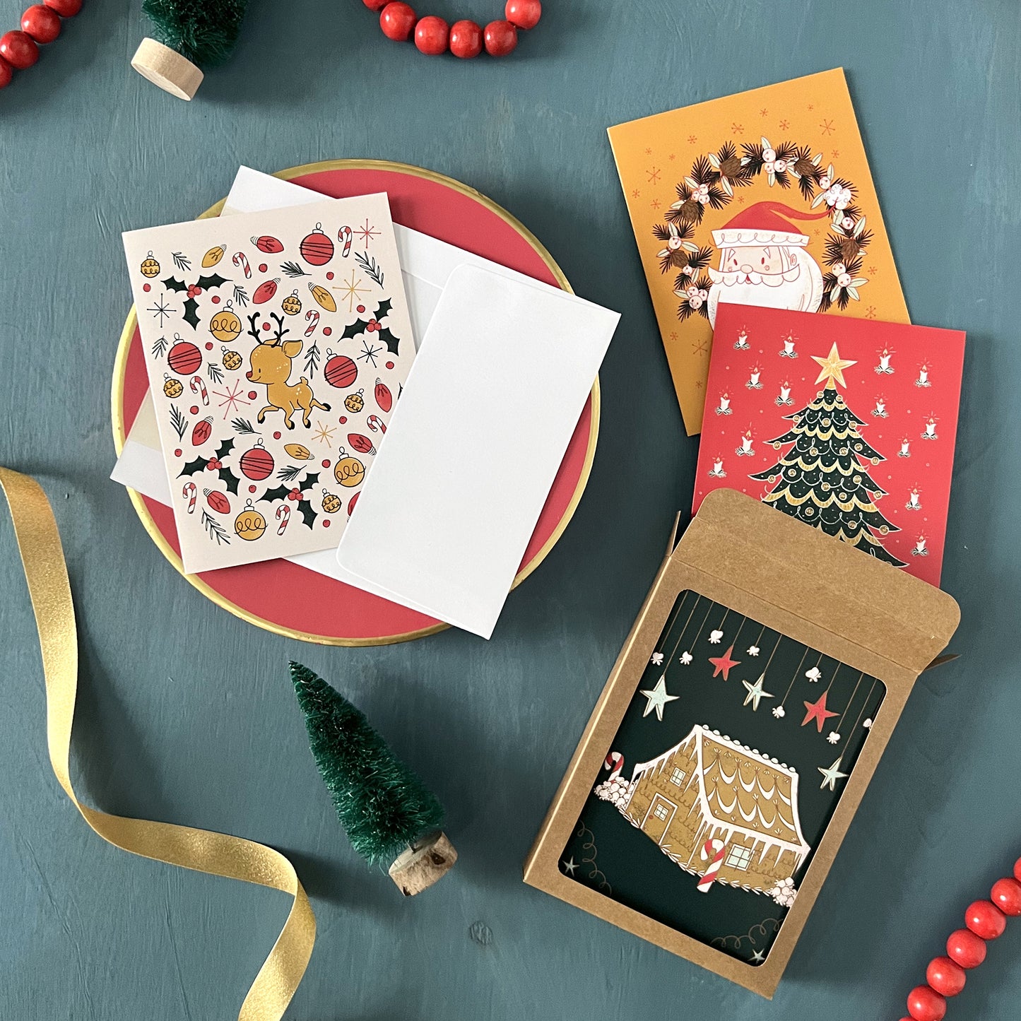 A boxed collection of 4 retro Christmas card designs appears against a festive blue background with bottle brush trees, red beads, and gold ribbon.