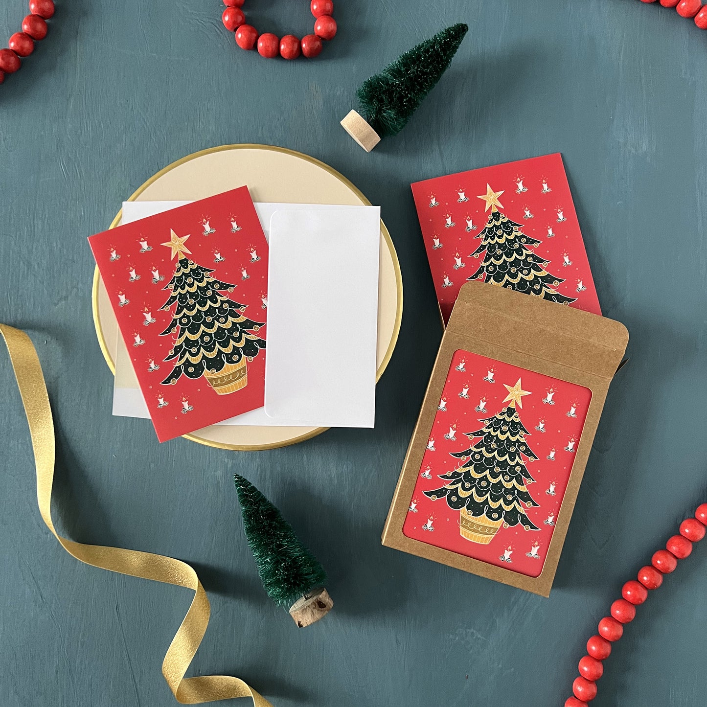 Red holiday greeting cards with retro Christmas trees appear on a festive blue background with red beads, green ribbon, and bottle brush trees.